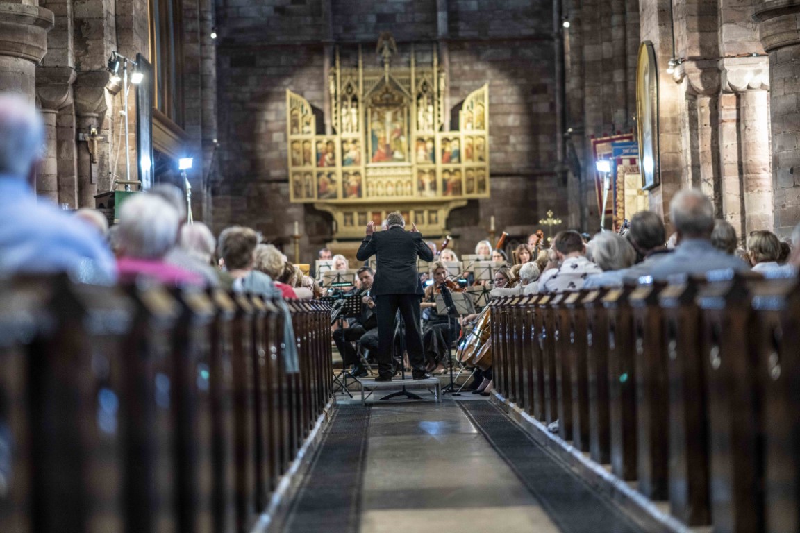 A view of the orchestra taken at a low level from the back of the Abbey along the main aisle and showing a packed audience. At the far end John Moore is conducting the orchestra at the chancel end. Credit Andy Bell Credit Andy Bell