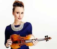 Jennifer Pike holding her violin. She has her hair tied up and is wearing a dark blue top with a pearl necklace